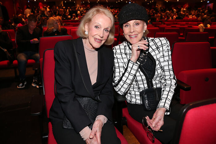 Beloved twin sisters, former entertainment icons, seated together in a theater with red seats and audience in background Beloved twin sisters, former entertainment icons, seated together in a theater with red seats and audience in background