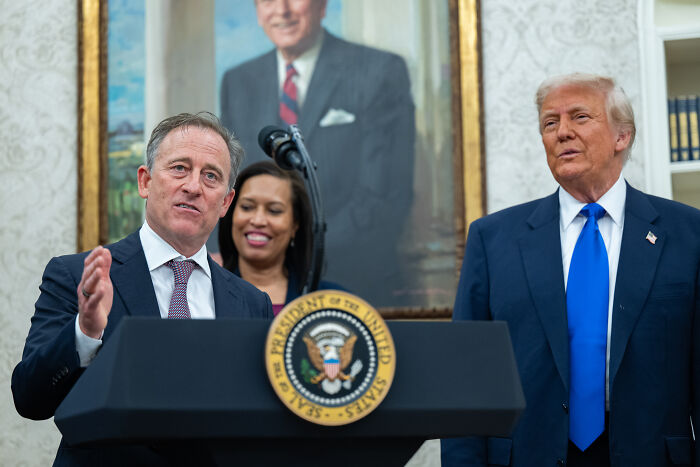 Former President Trump standing beside a speaker at a podium with the presidential seal inside a formal room.