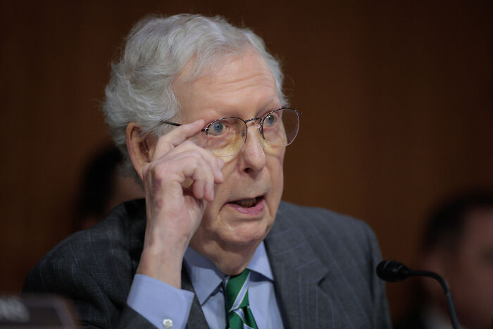 Elderly man in glasses speaking seriously at a hearing, related to government impact on family income. Elderly man in glasses speaking seriously at a hearing, related to government impact on family income.
