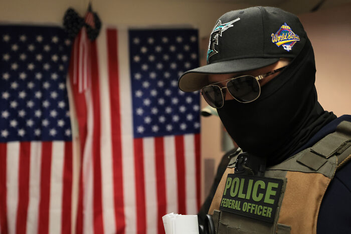 Federal officer in tactical gear and sunglasses with American flags in the background related to ICE nearly deporting Native American woman