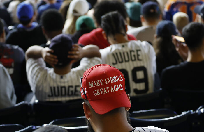 Person wearing a red Make America Great Again hat sitting among a crowd during a public event or gathering.