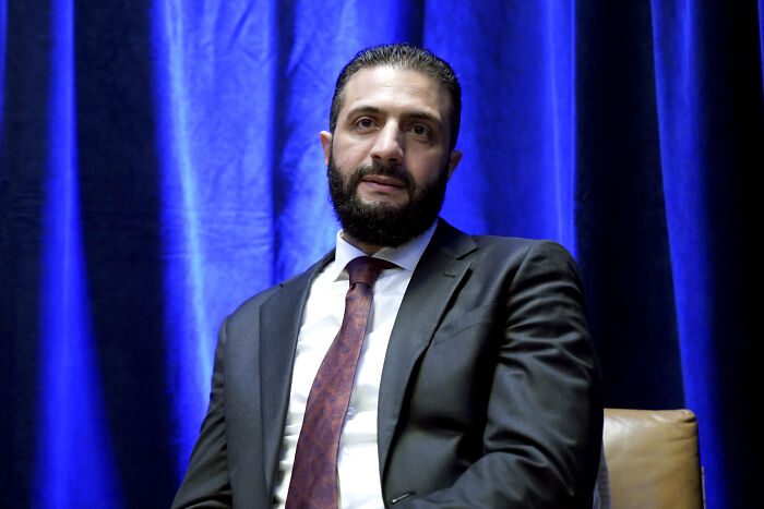 Man in suit and tie seated against blue curtain backdrop during White House meeting with Syrian president. Man in suit and tie seated against blue curtain backdrop during White House meeting with Syrian president.