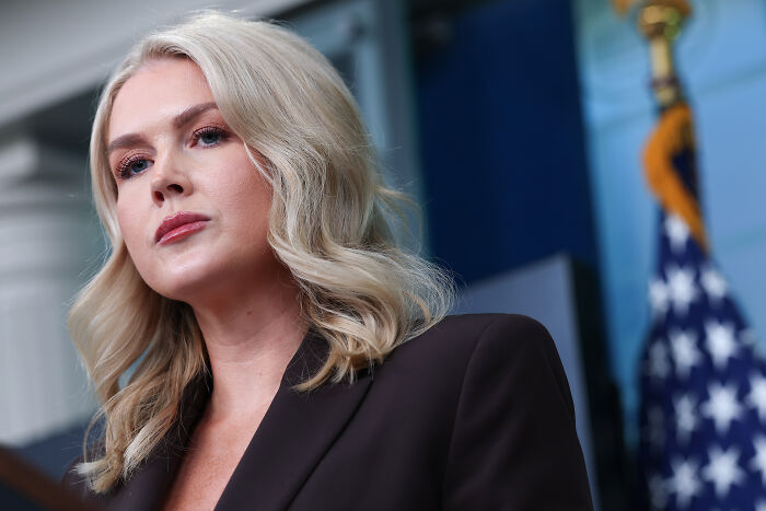Karoline Leavitt speaking at a podium, wearing a dark blazer with an American flag in the background. Karoline Leavitt speaking at a podium, wearing a dark blazer with an American flag in the background.