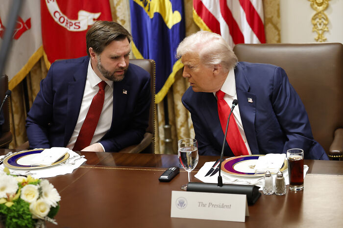 Donald Trump and JD Vance in a private conversation at a formal event with flags and a presidential nameplate visible. Donald Trump and JD Vance in a private conversation at a formal event with flags and a presidential nameplate visible.
