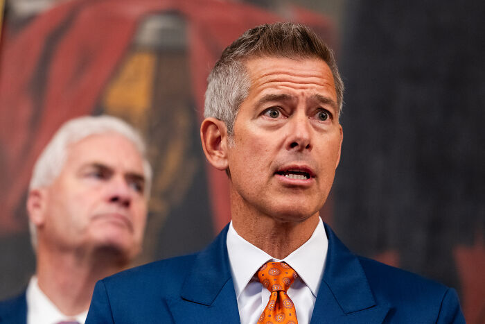 Sean Duffy speaking at a press event, wearing a blue suit and orange tie, responding to Gavin Newsom's photo.