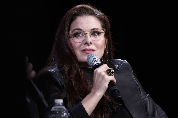 Debra Messing speaking into a microphone during an event, wearing glasses and a black leather jacket. Debra Messing speaking into a microphone during an event, wearing glasses and a black leather jacket.