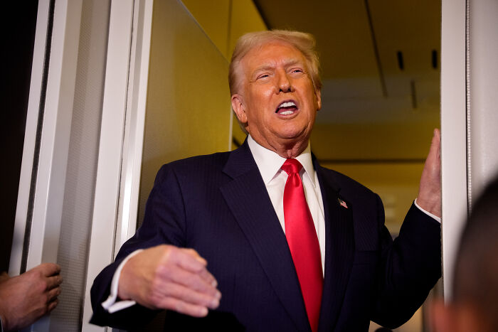 Donald Trump speaking indoors, wearing a dark suit and red tie, gesturing with his right hand near a doorway.