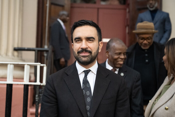 Zohran Mamdani wearing a suit, standing outdoors with people in background at a public event. Zohran Mamdani wearing a suit, standing outdoors with people in background at a public event.