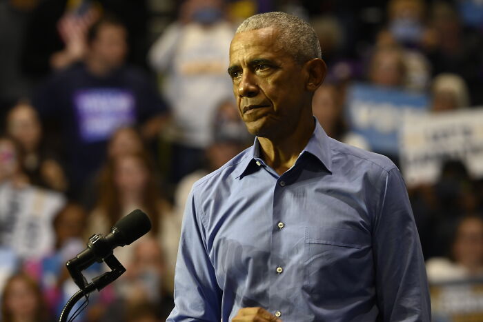 Barack Obama speaking at an event, wearing a blue shirt, roasting Donald Trump over costly White House makeover.