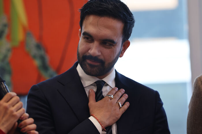 Zohran Mamdani wearing a dark suit, touching his chest thoughtfully during a discussion about democratic socialism. Zohran Mamdani wearing a dark suit, touching his chest thoughtfully during a discussion about democratic socialism.