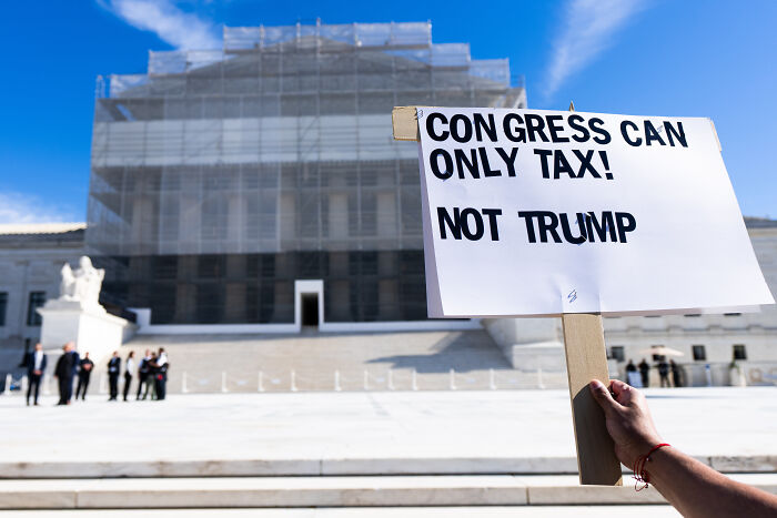 Protester holding sign about Trump tax repayments in front of a government building on a clear day.