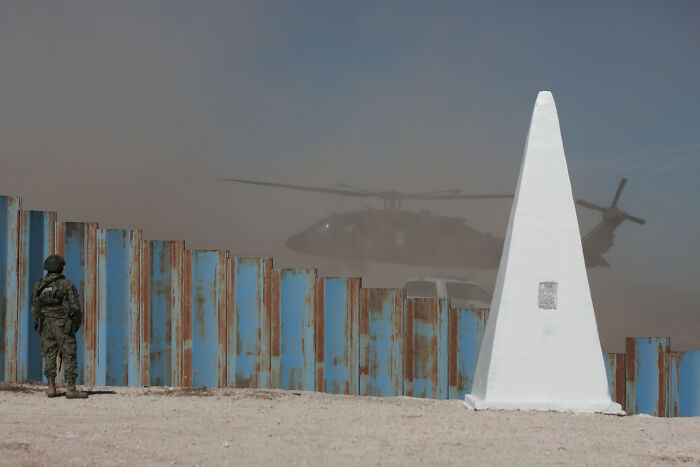 Soldier standing by a rusted border fence with a helicopter landing nearby, illustrating migration control and surveillance states. Soldier standing by a rusted border fence with a helicopter landing nearby, illustrating migration control and surveillance states.