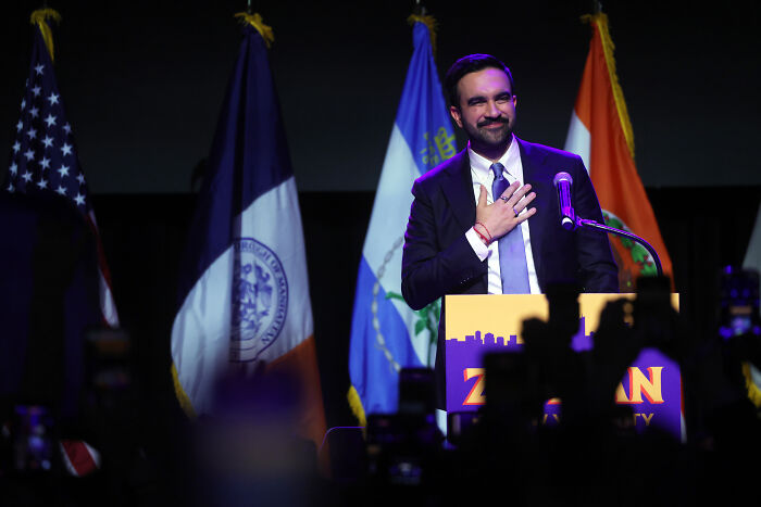 Zohran Mamdani at a podium with flags behind him, addressing audience after winning NYC mayoral race. Zohran Mamdani at a podium with flags behind him, addressing audience after winning NYC mayoral race.