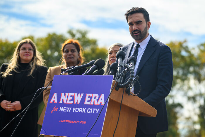 Zohran Mamdani speaking at a podium with a New York City sign, facing media and supporters outdoors. Zohran Mamdani speaking at a podium with a New York City sign, facing media and supporters outdoors.