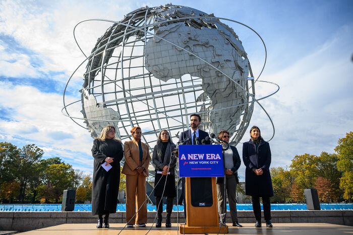 Zohran Mamdani speaking at a podium with a globe sculpture behind during NYC mayoral race event. Zohran Mamdani speaking at a podium with a globe sculpture behind during NYC mayoral race event.