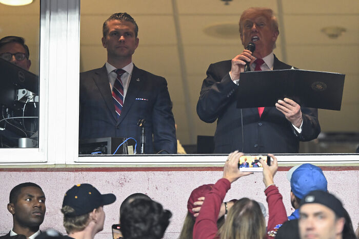 Trump speaking at a football game box while fans react with loud boos after stadium naming call.