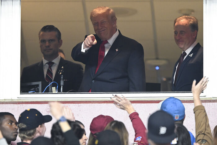 Donald Trump pointing from a box at a football game, reacting to loud boos from the crowd after stadium naming remarks.