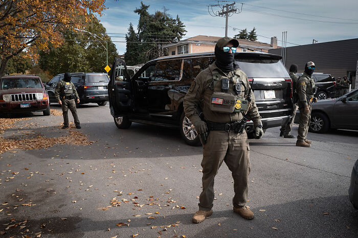 Federal agent wearing tactical gear standing near black SUV on a street during operation in Chicago neighborhood. Federal agent wearing tactical gear standing near black SUV on a street during operation in Chicago neighborhood.