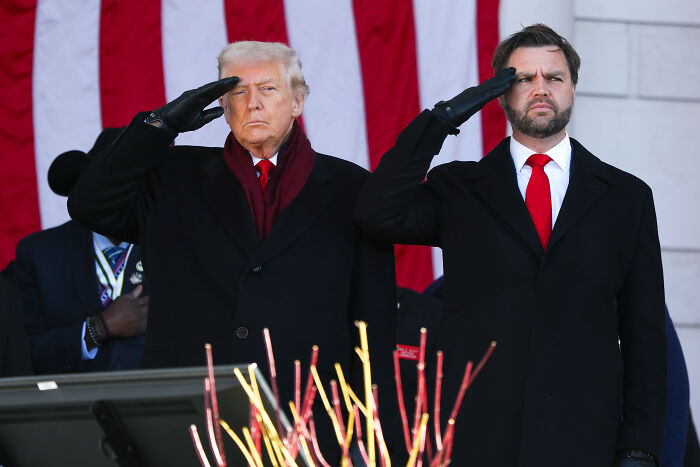 Donald Trump and another man saluting during a Veterans Day event with American flag backdrop. Donald Trump and another man saluting during a Veterans Day event with American flag backdrop.