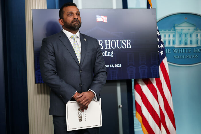 Kash Patel standing at White House briefing room in a suit, holding papers with an American flag nearby.
