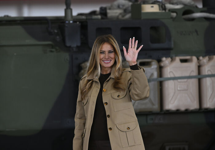 Melania Trump in a tan jacket waving to troops during a military event sparking public backlash over her dystopian address.