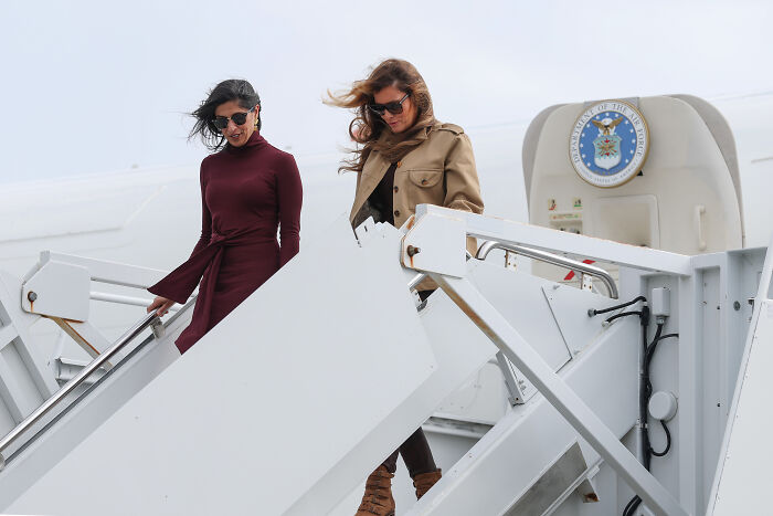 Two women wearing sunglasses and coats descend airplane stairs, with a military seal visible on the aircraft side.