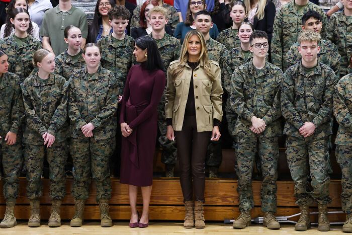 Melania Trump standing with troops and civilians during a public event, wearing a beige jacket and brown pants.