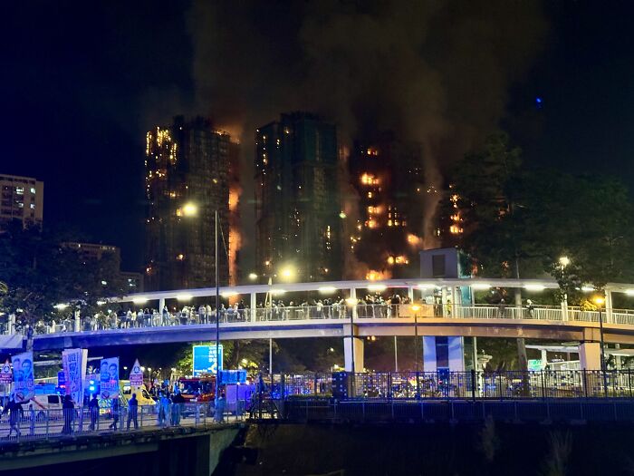 Hong Kong apartment complex fire at night with heavy flames and smoke, emergency responders and crowd on nearby bridge.