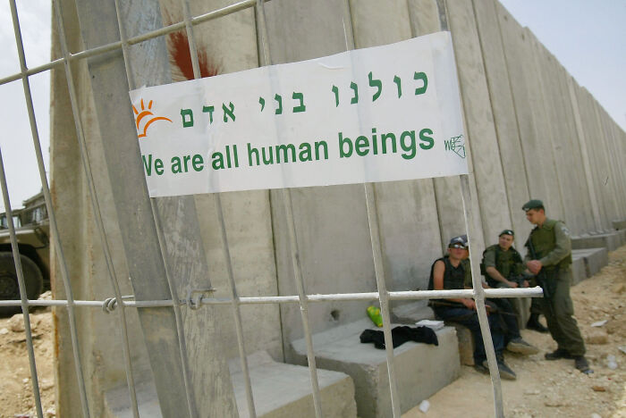 Banner on metal fence reads We are all human beings near concrete barrier with soldiers, illustrating migration control and surveillance states. Banner on metal fence reads We are all human beings near concrete barrier with soldiers, illustrating migration control and surveillance states.