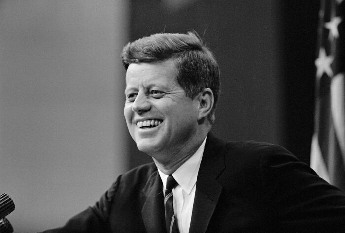 Black and white photo of President Kennedy smiling and speaking at a podium with an American flag in the background.