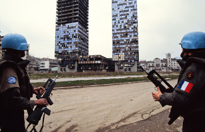 Two armed peacekeepers in blue helmets patrol a damaged urban area linked to human safari trips investigation.