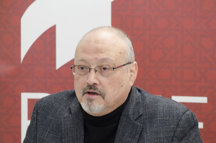 Jamal Khashoggi speaking at an event, wearing glasses and a dark blazer with a patterned red background. Jamal Khashoggi speaking at an event, wearing glasses and a dark blazer with a patterned red background.