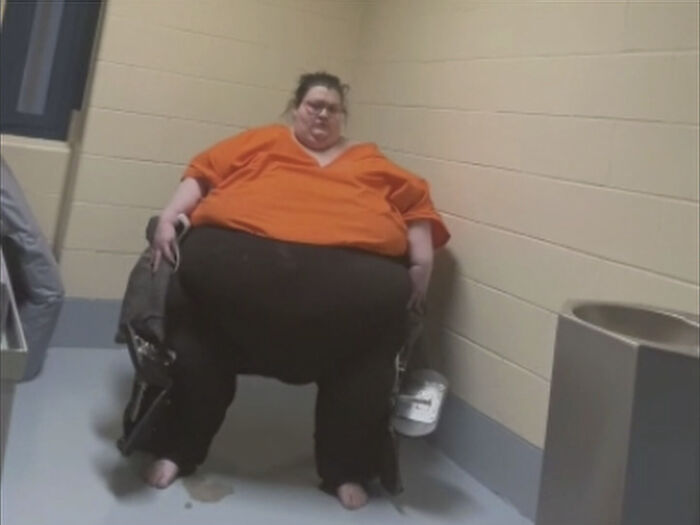 Obese woman in an orange shirt sitting against a beige wall in a confined room holding a small bucket and a tray.