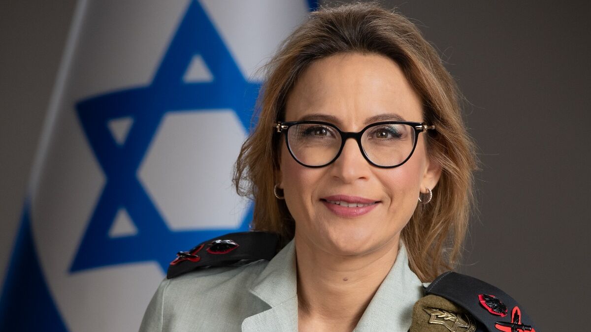 Israeli lawyer in military uniform with glasses, smiling in front of an Israeli flag background.