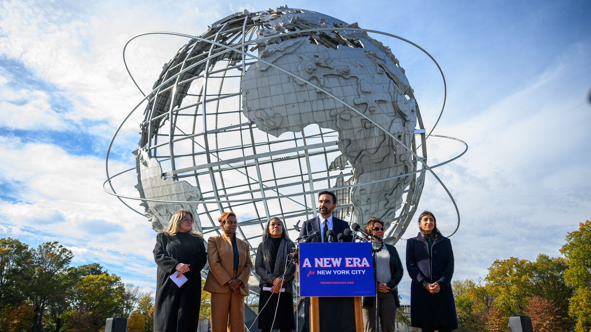 Zohran Mamdani speaking at press event with supporters in front of large globe sculpture in New York City park.