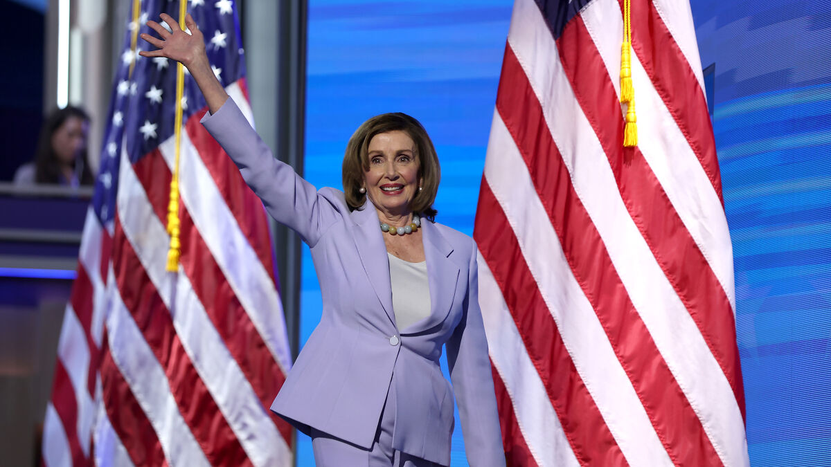 Nancy Pelosi in a lavender suit waving on stage with American flags, symbolizing her congressional career announcement.