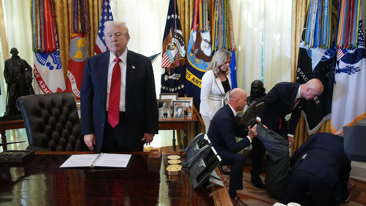 Donald Trump standing at a desk with a serious expression as a man faints behind him in an official room.
