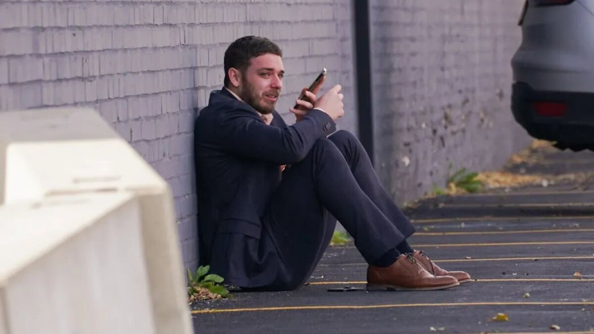 Man in tears sitting against wall holding phone, emotional moment near ICE facility parking lot.