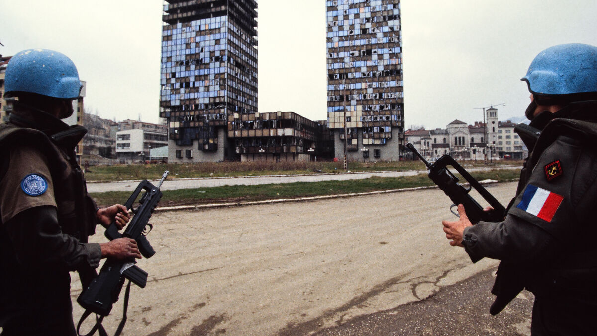 Two armed soldiers in blue helmets stand in a damaged urban area related to human safari trips investigation.