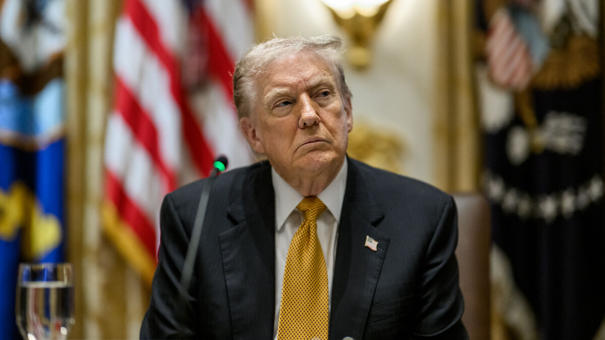 Donald Trump in a formal setting, wearing a dark suit and yellow tie, with American flags blurred in the background.