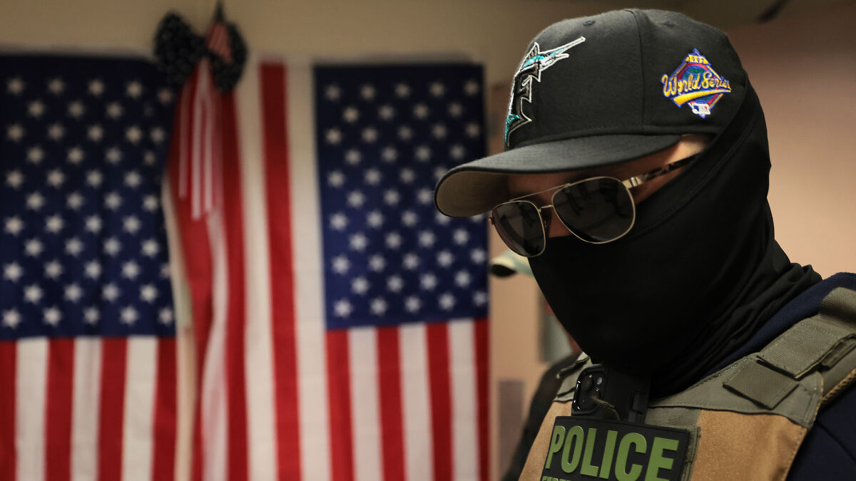 Law enforcement agent wearing sunglasses and tactical gear in front of an American flag, related to ICE deportation case.