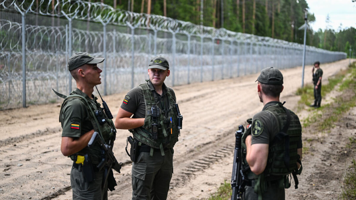 Border security officers standing near a fence with barbed wire illustrating migration control and surveillance states.