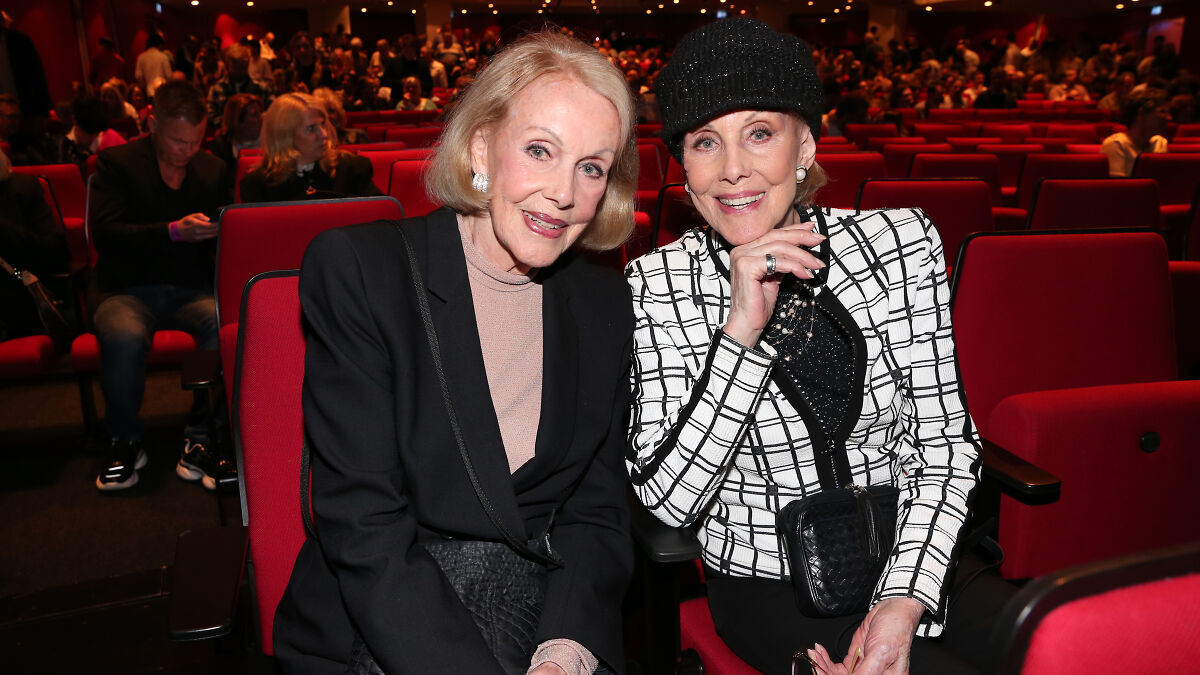 Twin sisters once entertainment icons smiling together seated in a crowded theater event setting.