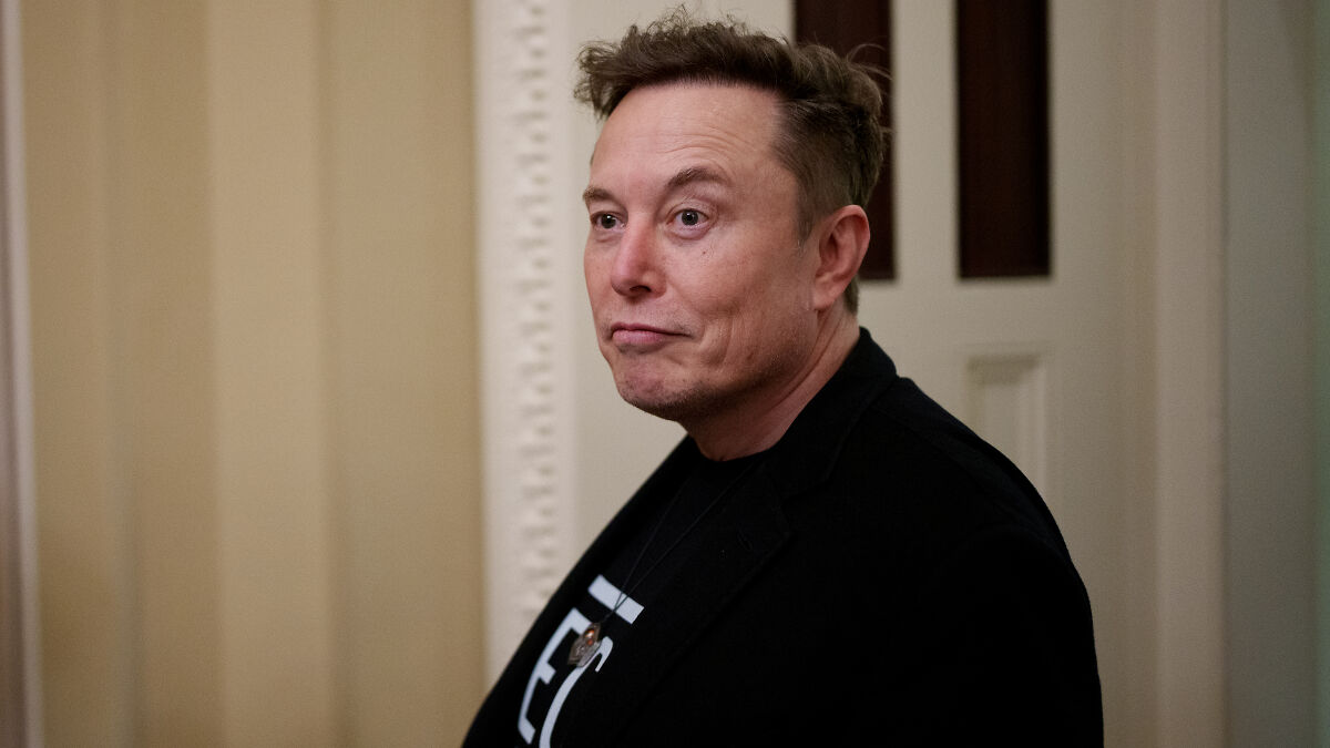 Elon Musk indoors wearing a black jacket with a sarcastic expression related to MAGA influencer accounts and foreign trolls.