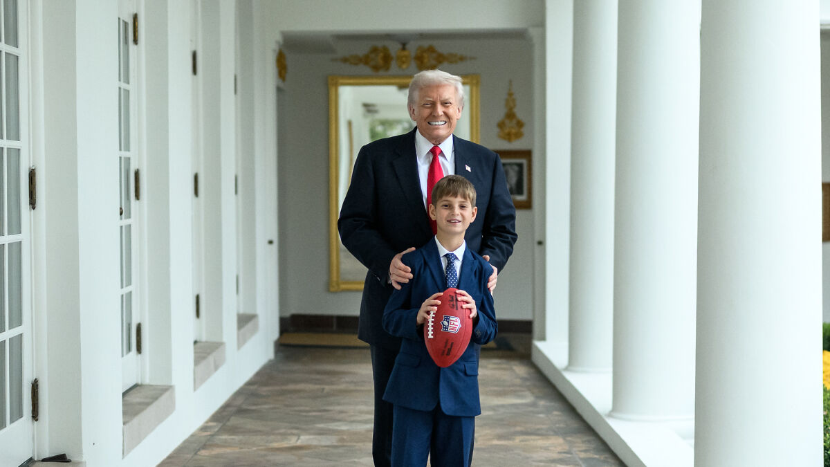 Donald Trump smiling and standing behind his grandson holding a football in a corridor with white columns.