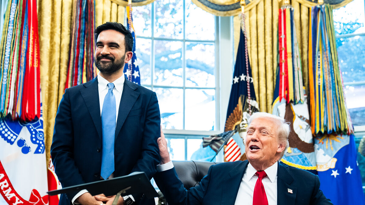 Zohran Mamdani smiling in the White House with former president, surrounded by flags and large windows behind.