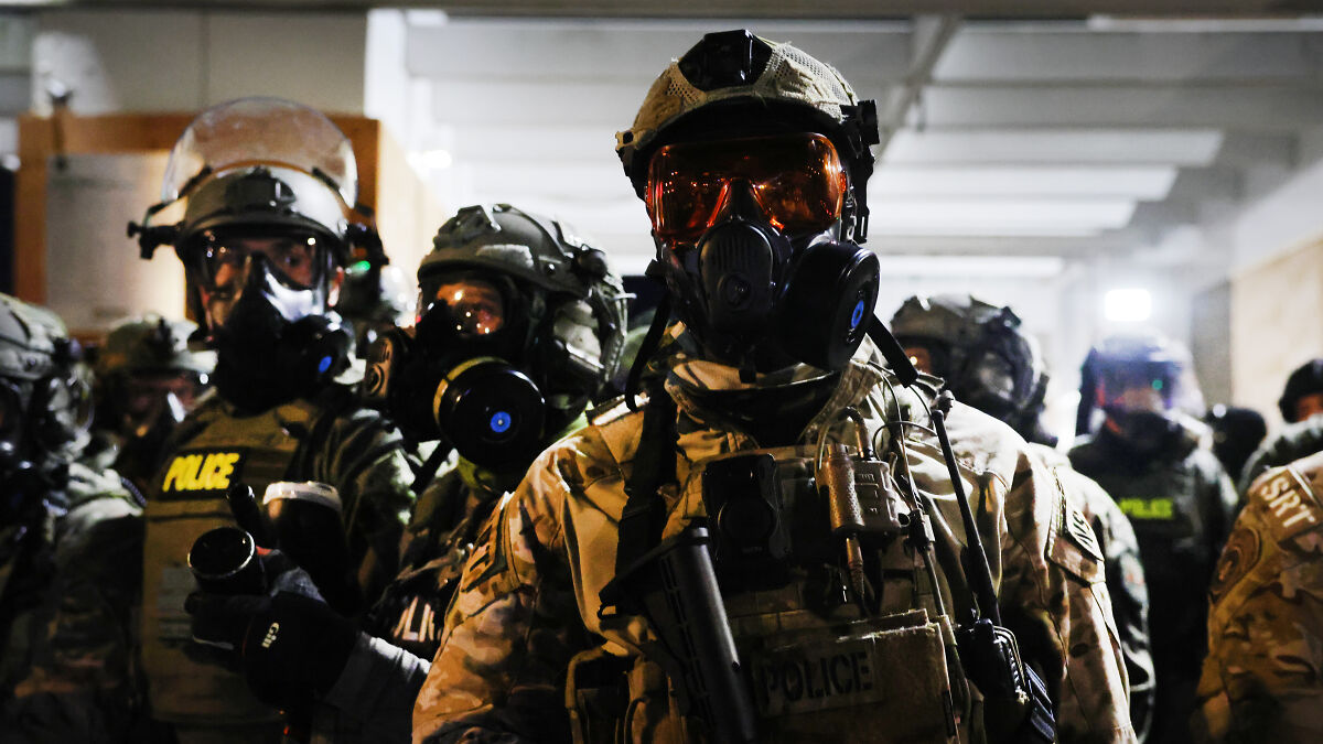 Heavily armed police officers in tactical gear and gas masks preparing for a high-risk ICE raid operation indoors.