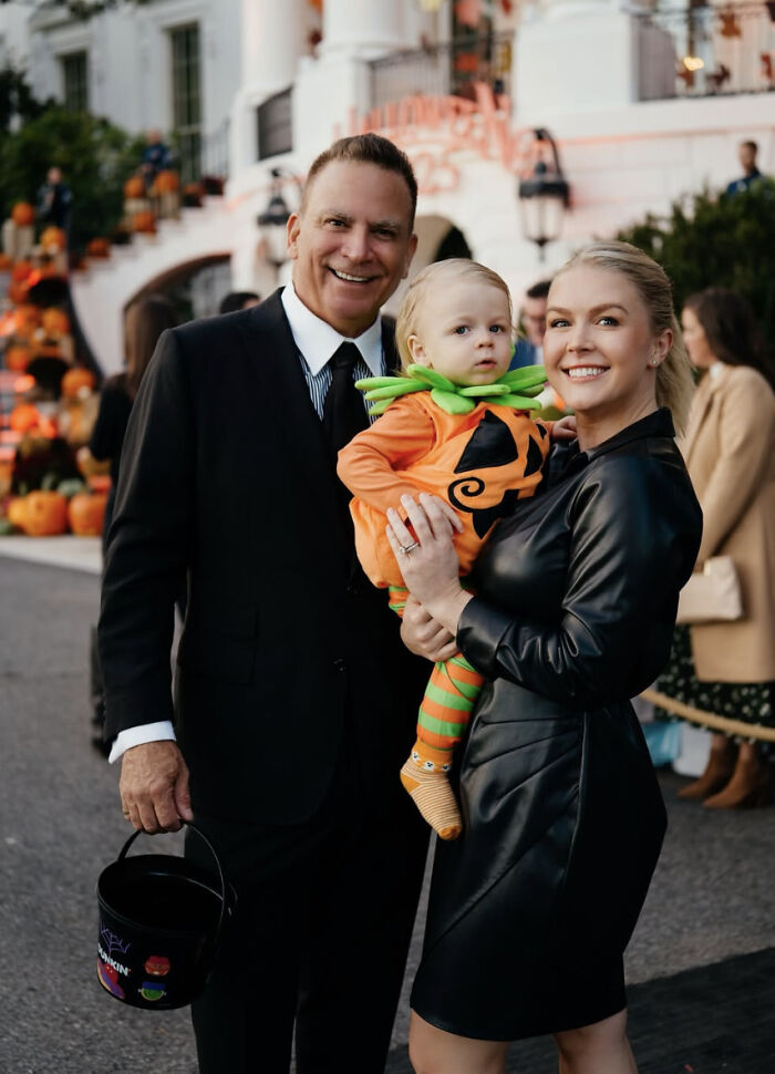 Karoline Leavitt smiling in black dress holding child in pumpkin costume with husband in suit during Halloween event. Karoline Leavitt smiling in black dress holding child in pumpkin costume with husband in suit during Halloween event.