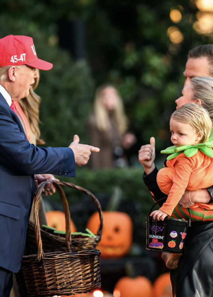 Man in red hat handing out Halloween treats while woman holds child in pumpkin costume during outdoor event Man in red hat handing out Halloween treats while woman holds child in pumpkin costume during outdoor event