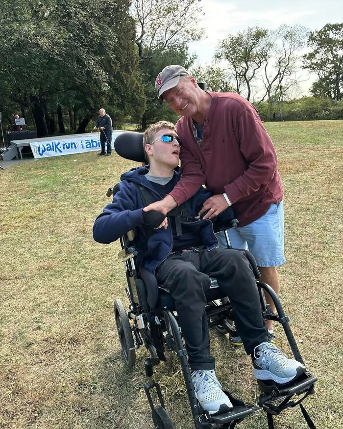 Man in cap smiling and holding hands with his disabled son in a wheelchair outdoors at a walk/run event.